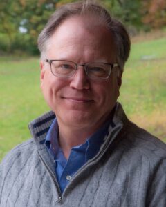Image of white male with glasses wearing a grey sweater and blue button up shirt. 
