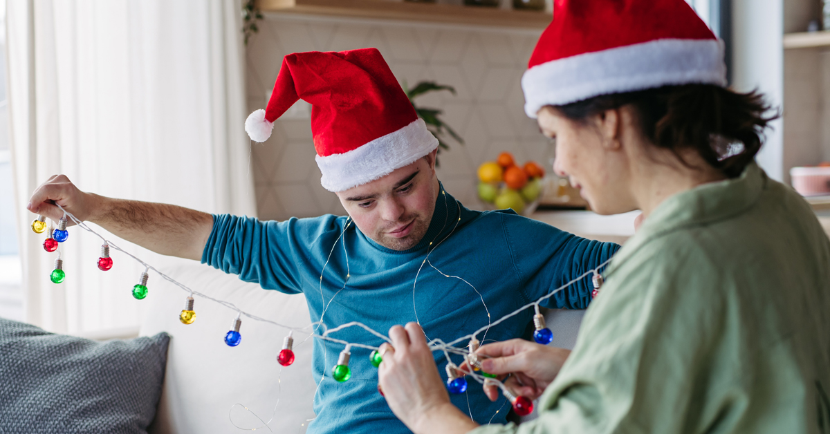 Two people sit in a living room wearing Santa hats while detangling holiday lights.