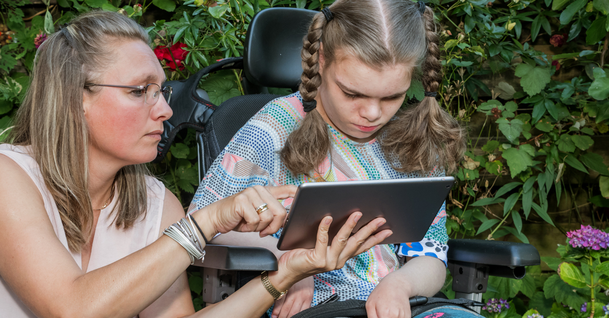 A white, female direct support professional assist a younger white woman, who has a disability and uses a wheelchair, with operating a tablet device for communication