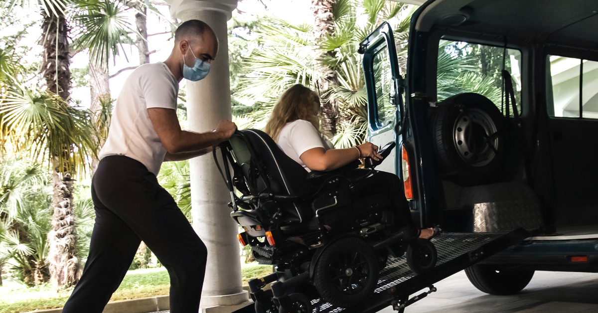 A direct support professional assists a woman using a wheelchair to enter a van, with a ramp.