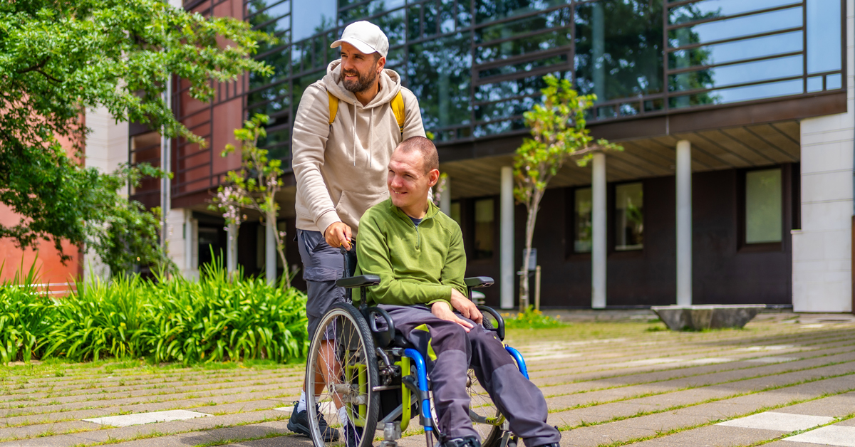 One man supports another man who is using a wheelchair. They are outside  in a park-like setting on a summer day.