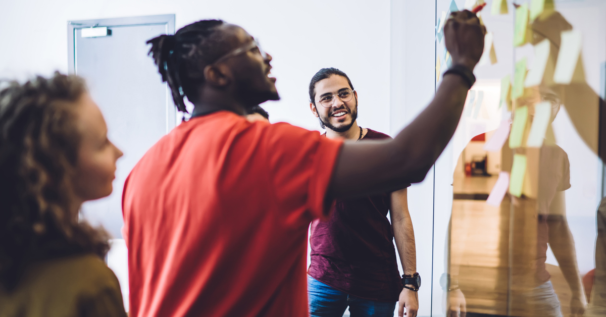 A black man wearing a red shirt points his pen at a whiteboard with sticky notes. Two other colleagues observe.